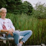 Mo Kenney with blonde hair sitting in an folable chair in a field of long grass in Nova Scotia wearing white tee and light blue jeans