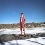Mo Kenney press image of Mo in Nova Scotia standing on a wooden walkway wearing a peach trouser suit and white shirt looking confident