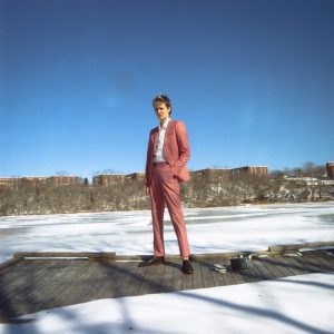 Mo Kenney press image of Mo in Nova Scotia standing on a wooden walkway wearing a peach trouser suit and white shirt looking confident for Ahead of Myself album promo